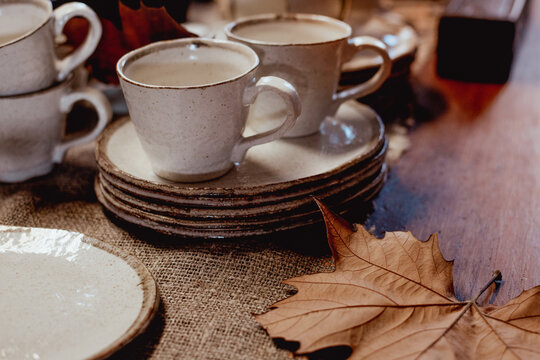 Two ceramic cups sit on stacked plates atop a burlap surface, accompanied by a dried leaf
