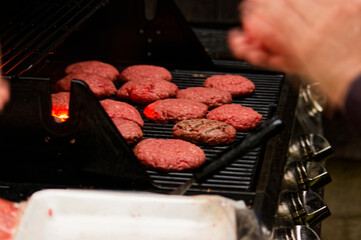 Beef patties sizzle on hot outdoor grill showing visible grill marks, one slightly charred. Hands use spatula to cook burgers, perfect for restaurants or catering events.