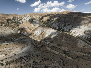 Aerial view of stark, sun-drenched mountains reveal contrasting hues of earth and stone, bisected by a winding road, Vardenyats Pass, Vayots Dzor, Armenia.