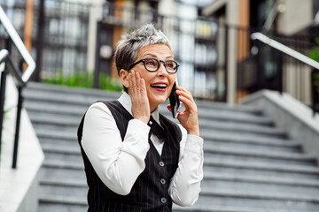 Mature businesswoman conversing on a smartphone outdoors with a joyful expression in an urban...