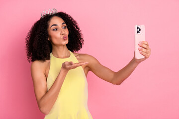 Young woman in yellow dress taking a selfie on pink background with a crown, expressing lively and...