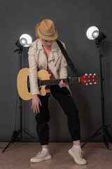 Expression female musician plucking classical guitar, looking down at neck of acoustic guitar. Playful woman guitarist standing full length on stage, dressed in leather jacket, straw hat, jeans pants