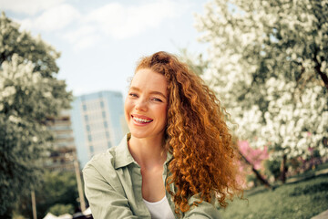 Charming young woman with red curly hair enjoying a sunny day outdoors in a blooming park during...