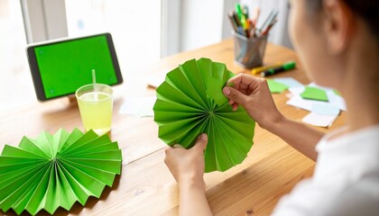 A person's hands meticulously crafting a vibrant green paper decoration on a wooden desk, with a tablet displaying a green screen nearby.