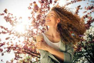 Young woman enjoying a beautiful spring day under blooming trees with red curly hair and a charming...