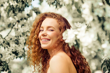 Attractive young woman with red curly hair enjoying a sunny day in a blooming park, exuding beauty...