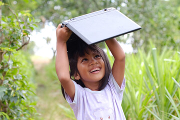 a young girl with black hair is holding a laptop. She is standing outdoors amidst lush greenery. The light-colored shirt she is wearing has a small design. Her expression is happy and natural smile.