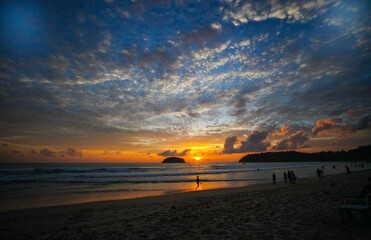 Golden Embrace above Kata beach.
The setting sun paints the horizon with golden light, reflecting across gentle waves as silhouettes of beachgoers stroll peacefully along the shoreline.