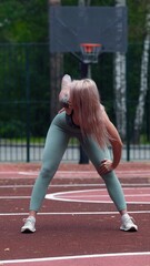 Young blonde woman in sportswear doing stretching exercises standing on basketball court in the...