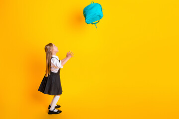 Cheerful schoolgirl in uniform catching backpack on vibrant yellow background, embodying excitement