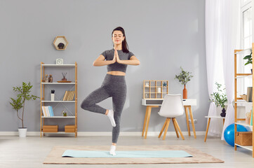 Sporty slim young woman practicing yoga standing in Vrksasana position on sport mat at home. Happy...