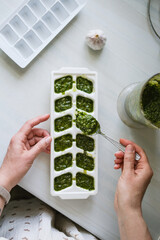 Vertical top-down view of female hands filling an ice cube tray with green pesto using spoon. On...