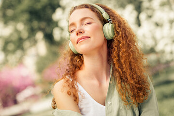 Lovely woman with curly red hair enjoying music with headphones while relaxing outdoors during a...