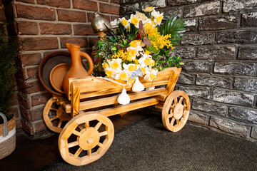 A decorative wooden cart with carved wheels, filled with bright flowers, decorated with a clay jug...