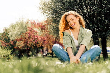 Happy young woman with curly red hair enjoying a sunny day outdoors in a peaceful park during...