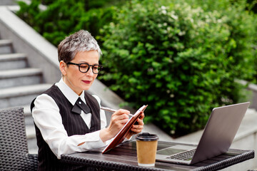 Confident businesswoman working outdoors in an urban park with laptop and clipboard, showcasing...