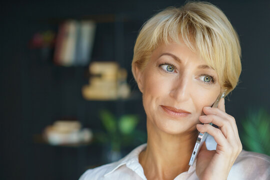 Caucasian woman, 40-50 years old, engages in a serious conversation on her mobile phone indoors. Bookshelf and plants create a thoughtful atmosphere in the home office setting.