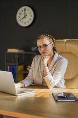 Caucasian woman, 25 years old, sits focused at her laptop in a bright office. Serious expression reflects confidence as she balances her smartphone and tablet.