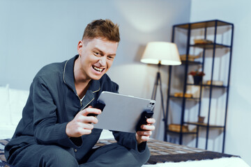 Young man joyfully plays video game on tablet using controllers in cozy bedroom. Evening light creates a relaxed atmosphere filled with playfulness and excitement.