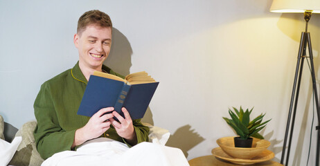 Happy young man immersed in reading a book in a cozy bedroom. Soft lighting from the floor lamp creates a peaceful atmosphere, enhancing the calm evening mood.