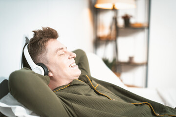 Happy young man enjoys peaceful moment in cozy bedroom while wearing headphones. Sunlight filters through the window, creating a relaxed atmosphere.