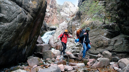 A guide and a tourist are walking along a rocky canyon, with a small waterfall visible behind them. Two friends are hiking in the mountains