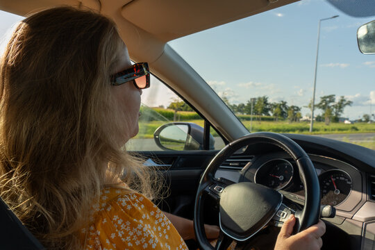 Female driver in stylish sunglasses enjoying peaceful countryside drive with rural landscape and blue sky visible outside. Concept of leisurely travel, driving safety and summer road trip exploration.