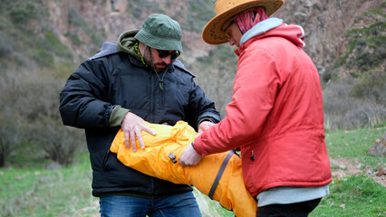 Two travelers are stuffing the tent into a cover. Two men are standing in a field and breaking...