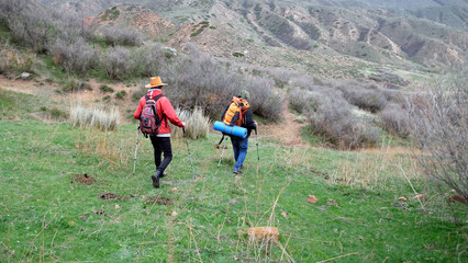 Two brothers are walking through a field with backpacks, and one of them is wearing a red jacket and a funny cowboy hat. The guys are walking through the wilderness