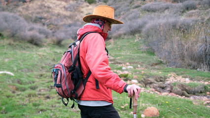 A lonely wanderer travels through the mountains. A young guy in a cowboy hat, backpack, trekking poles and yellow glasses walks through a green field
