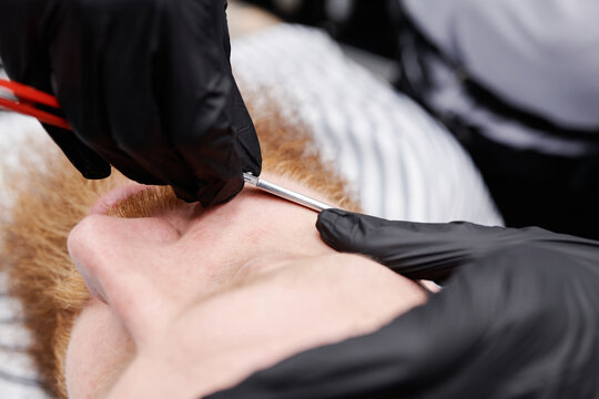 Horizontal close-up shot of unrecognizable barber wearing protective latex gloves working in barbershop shaving mans beard using razor blade