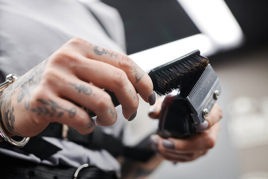 Low angle view close-up shot of unrecognizable professional hairdresser with tattooed hands cleaning hair clipper wiping off hair