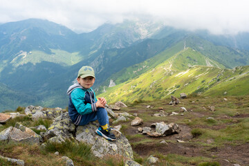 Fototapeta premium Happy young boy in cap and blue jacket sitting on rocky mountain boulder enjoying spectacular alpine valley views on clear day. Concept of family hiking achievements and childhood mountain experiences