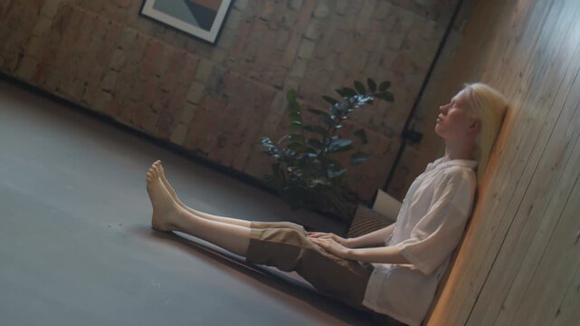 Unusual rotating shot of young woman with albinism wearing casual outfit lying on floor with eyes closed and legs up against wall, sitting on wall effect