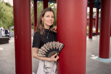 Portrait of a stylish woman in a black and white dress holding an ornate lace fan while leaning against a large red column in a traditional oriental pavilion