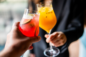 Two hands holding vibrant summer drinks—one red with lime slices, the other orange with pulp—against a blurred background. Refreshing beverages in a celebratory moment