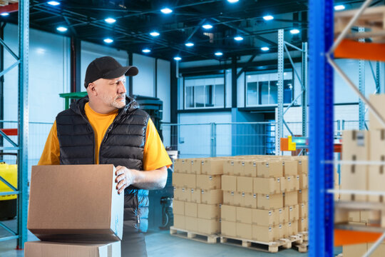 Man works in warehouse. Guy with box in fulfillment center building. Man inside logistics warehouse. Guy in storage area of industrial factory. Adult person works inside warehouse hangar.
