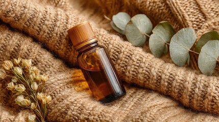 Amber glass bottle of essential oil, resting on a draped, beige knit sweater, surrounded by sprigs of eucalyptus and dried flowers