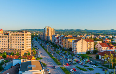  Evening cityscape of Novi Sad, Serbia, featuring Bulevar Europe lined with modern residential buildings. Warm sunlight casts long shadows over streets, cars, and distant green hills.