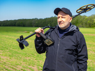 Smiling man holding metal detector on shoulder in green field under clear sky, enjoying outdoor treasure hunting. Metal Detecting, Archaeology, Outdoor Activity, Travel.