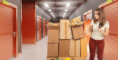 Woman with smartphone organizing cardboard boxes in a self-storage unit, pointing at labeled box with teddy bear. Activity: Moving, Storage, Relocation, Logistics, Self-storage.