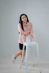 Young woman posing confidently beside a white bar stool in a minimalistic studio setting with soft...
