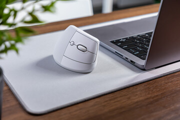 White ergonomic vertical mouse resting on a grey mouse pad next to a laptop, promoting comfort and...