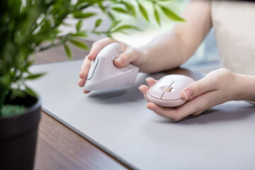 Person's hands compare a pink vertical ergonomic mouse and a standard wireless mouse on a grey pad,...