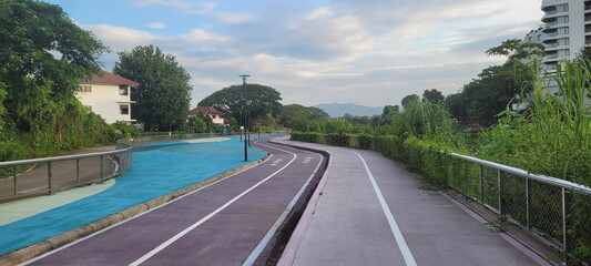 Quiet outdoor running track with multiple lanes, surrounded by greenery and residential buildings, under partly cloudy sky, creating peaceful and scenic environment for exercise and leisure