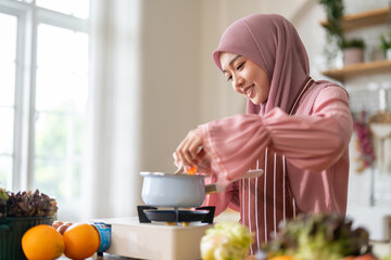 Muslim woman happy cooking healthy meal in kitchen wearing hijab making soup in pot with fresh...