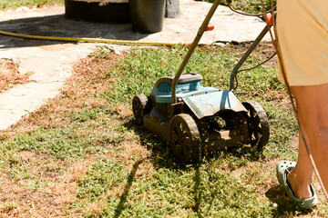 Blue and black lawn mower in action, cutting grass with clippings flying, path leading to a well,...