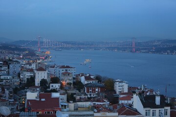 Obraz premium The illuminated Bosphorus Bridge connecting Europe and Asia at twilight, with boats crossing the strait and Istanbul’s city lights glowing.