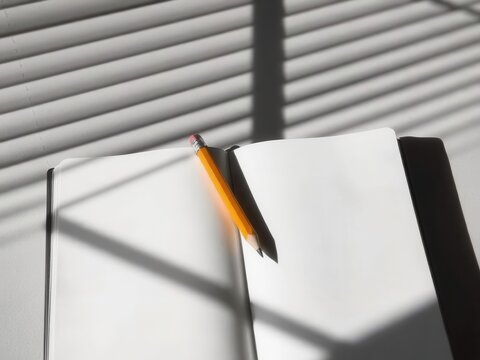 A blank notebook with a yellow pencil rests on a table. Soft shadows from blinds create a pattern on the surface. Ideal for Teacher's Day themes.