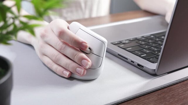 Close-up of a person's hand using a white ergonomic vertical mouse on a desk with a laptop, highlighting comfort and productivity in modern office work